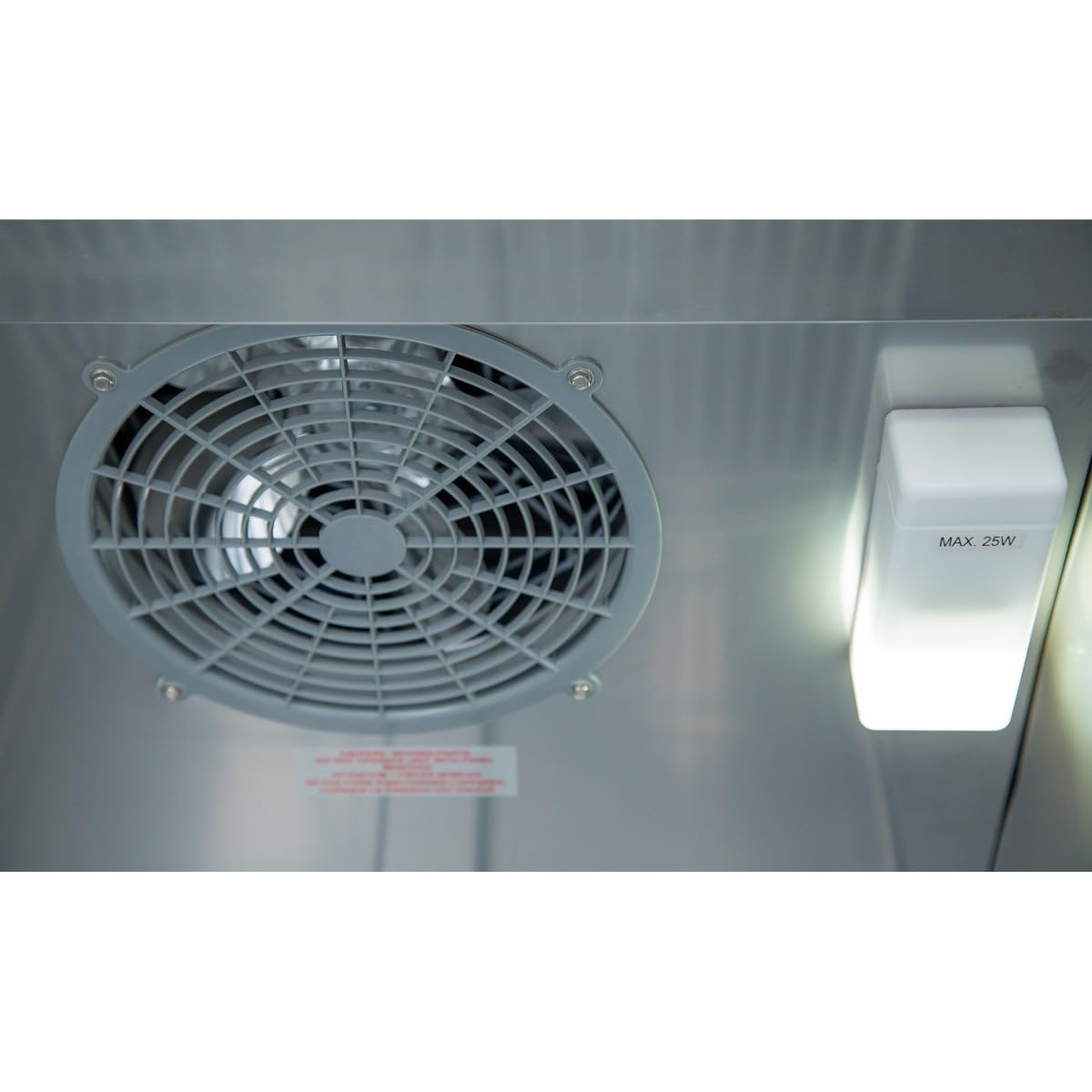 Close-up view of a ventilation fan with a circular grill next to a “MAX. 25W” light fixture inside a commercial refrigerator. The reflective surface and small red warning label suggest it’s likely a stainless steel refrigerator or reach-in model.