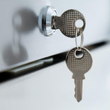 Close-up of two metallic keys on a keyring, with one key inserted into the lock of a shiny stainless steel refrigerator drawer. The other key hangs below. The reflective surface creates a clean, modern appearance.