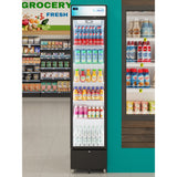 A tall, slim commercial merchandiser refrigerator in a grocery store displays an array of drinks—milk, juice, tea, and water. Groceries like canned goods and dairy products line nearby shelves with clear price tags. The store is clean and brightly lit.