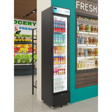 A tall black commercial merchandiser refrigerator with a glass door displays various drinks, including soda cans and bottled water, in a grocery store near shelves of packaged goods and a produce section with fresh fruits and vegetables.