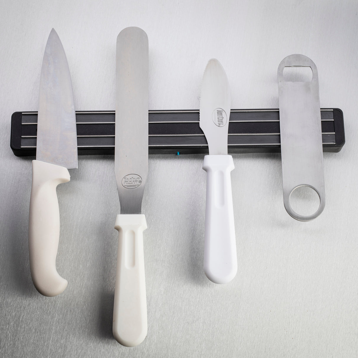 Four kitchen utensils, including two knives, an icing spatula, and a bottle opener with white handles, hang vertically from a black magnetic holder wall mount on a light grey textured surface. All items are dishwasher safe for easy cleaning.