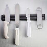 Four kitchen utensils, including two knives, an icing spatula, and a bottle opener with white handles, hang vertically from a black magnetic holder wall mount on a light grey textured surface. All items are dishwasher safe for easy cleaning.