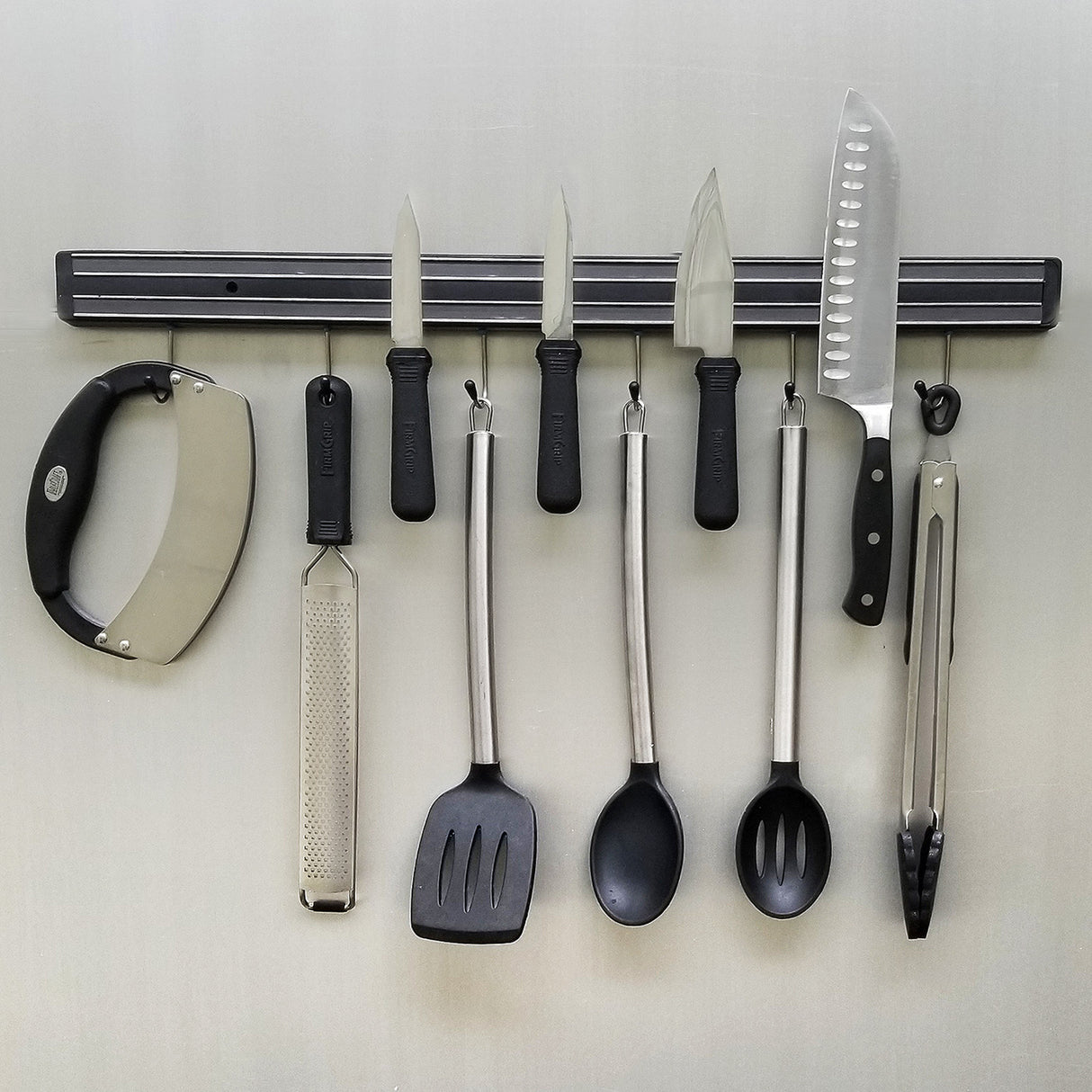 A wall-mounted magnetic holder keeps kitchen tools—mezzaluna chopper, grater, knives, spatula, spoons, and tongs—neatly arranged in a row, perfect for a cash & carry setup or organized home cooking space.