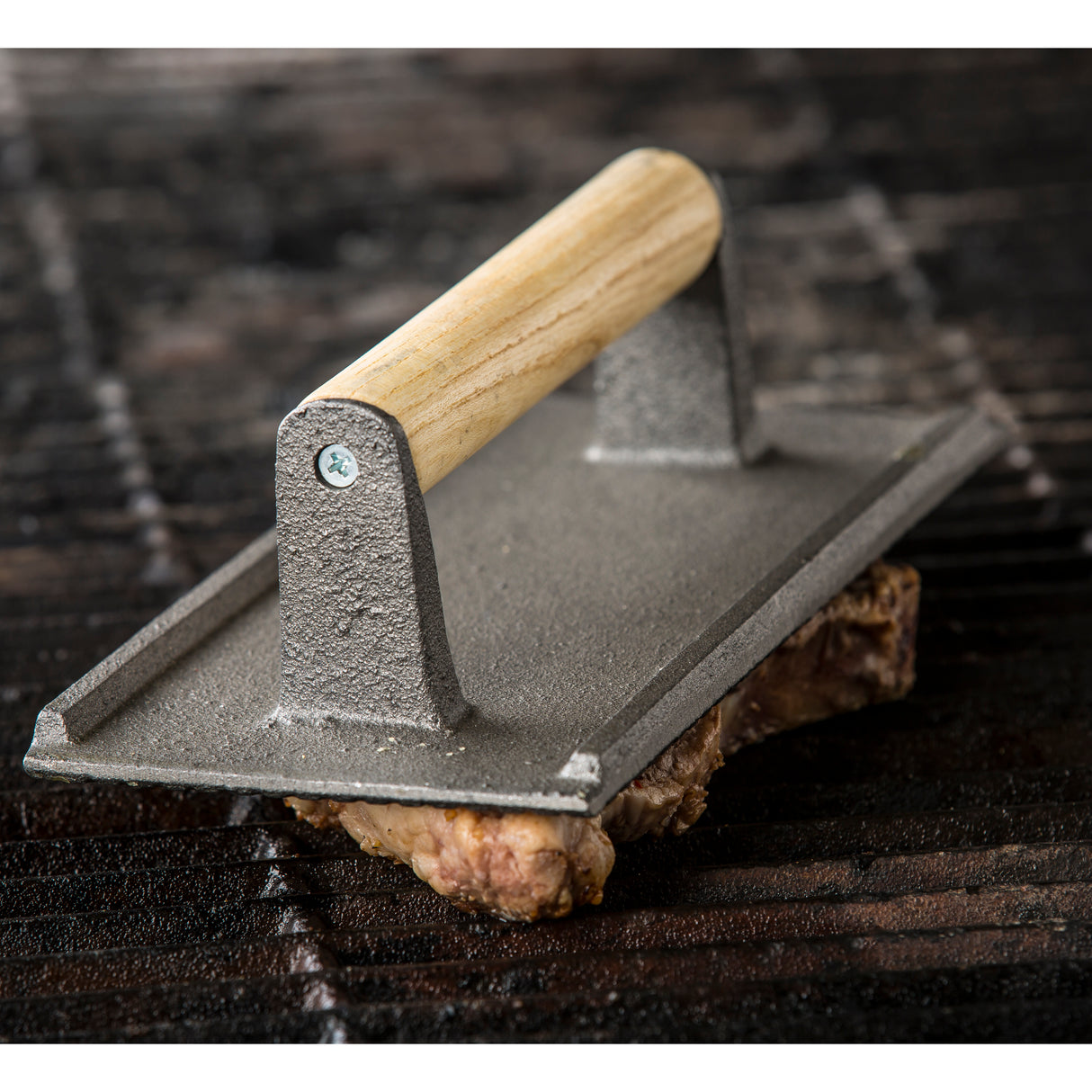 A heavy metal steak weight with a rectangular base and wood handle rests on a piece of meat, pressing it against dark grill grates. The surface has a rustic, textured finish, suggesting outdoor grilling or cooking.