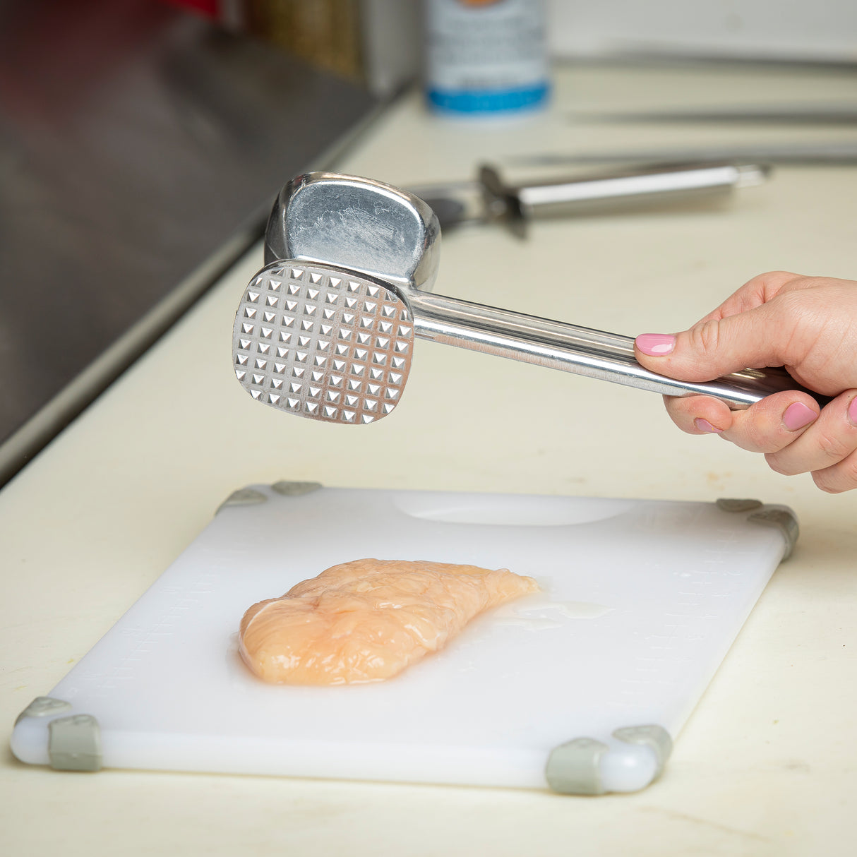 A hand holds a double sided cast aluminum meat tenderizer above a raw chicken breast on a white cutting board with gray corners; kitchen utensils and a spray can are visible in the blurred background.