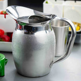 A shiny, textured Bell Water Pitcher with a wide spout and handle sits on a gray counter. In the background are blurred containers with assorted fruits, including strawberries and yellow pieces, along with a tall metal cup.