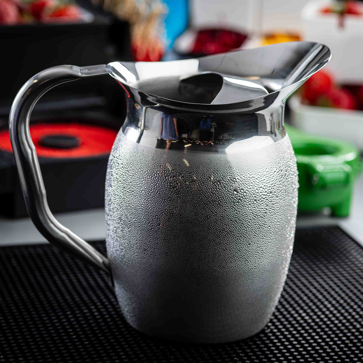 A stainless steel pitcher, possibly a 2 qt pitcher, is covered in condensation and placed on a black rubber mat. The blurred background features various colorful objects, including a green container and red items, evoking a casual kitchen-like atmosphere.