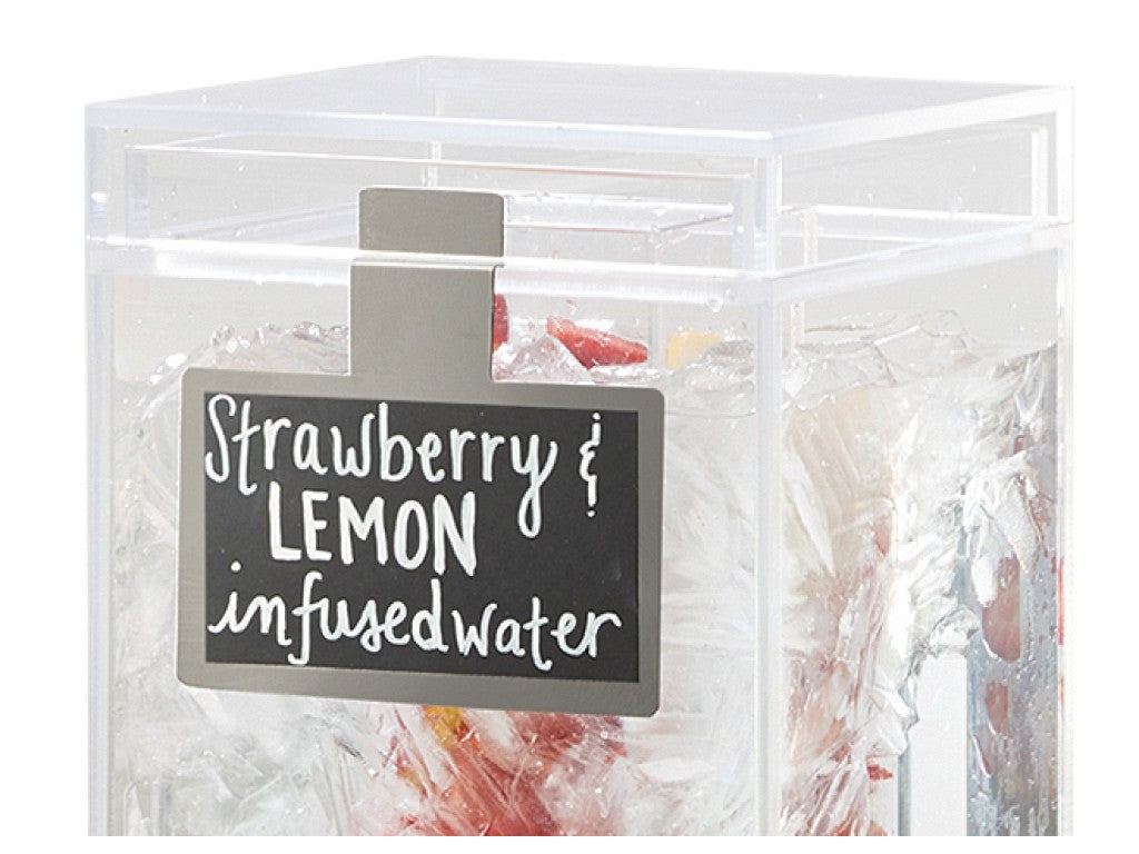 A clear square beverage dispenser filled with ice, sliced strawberries, and lemon pieces. A black write-on surface label clipped to the front reads “Strawberry & LEMON infused water” in white handwritten-style text. Only the upper part is visible.
