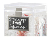 A clear square beverage dispenser filled with ice, sliced strawberries, and lemon pieces. A black write-on surface label clipped to the front reads “Strawberry & LEMON infused water” in white handwritten-style text. Only the upper part is visible.