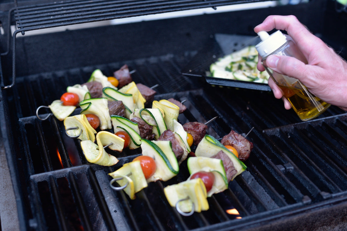 A hand sprays oil onto colorful vegetable and beef skewers grilling on a barbecue. The stainless steel skewers include zucchini, yellow squash, cherry tomatoes, and beef chunks. In the background, more sliced vegetables cook in a grill basket.