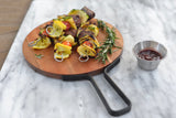 Three stainless steel skewers with grilled vegetables and meat are arranged on a round wooden cutting board with rosemary sprigs. A small metal cup of barbecue sauce sits nearby. Each 6 inch skewer holds yellow squash, zucchini, tomato, and beef cubes.