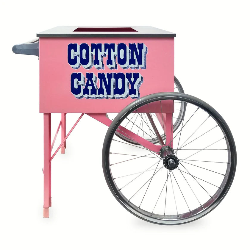 A Pink Floss Cart with large metal wheels and bold blue letters on the side that read COTTON CANDY. Made by Gold Medal Products, this floss unit cart has an open top, a left handle, and a charming vintage carnival feel.
