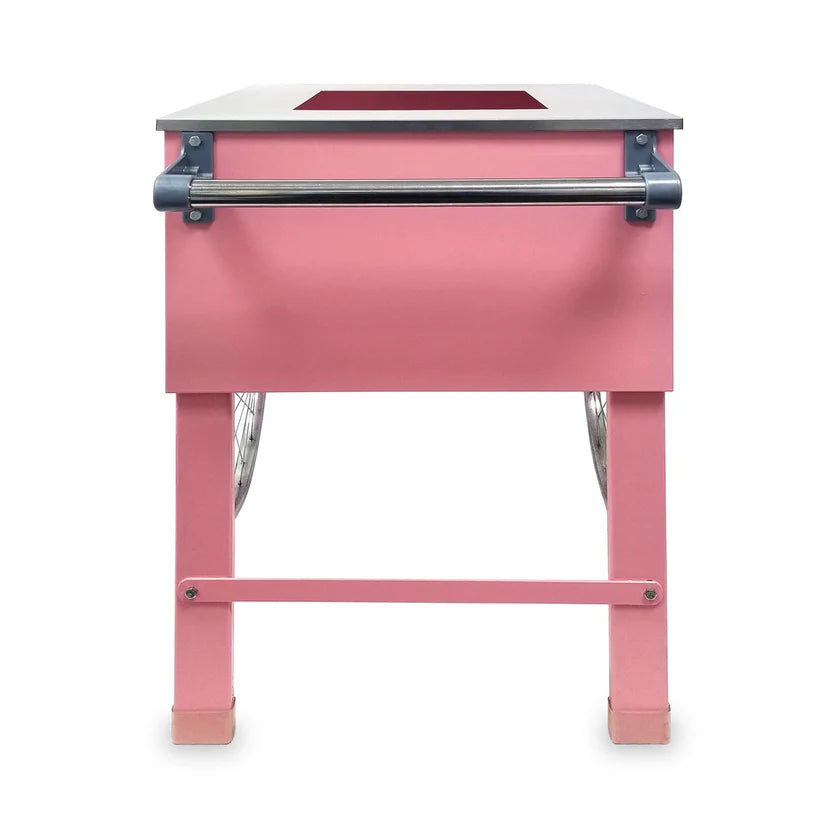 A pink rectangular Pink Floss Cart with four straight legs, a horizontal metal bar across the front, and visible metal wheels on each side. The tabletop has a rectangular cutout near the back edge. The background is plain white.