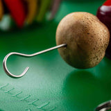 A stainless steel skewer pierces a whole mushroom, resting on a green textured surface. Out-of-focus, colorful vegetables appear in the background, suggesting food preparation for grilling or cooking.