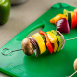 A stainless steel skewer holds sliced mushroom, red onion, yellow squash, and colorful peppers on a green cutting board, with another skewer and a green bell pepper blurred in the background. Dishwasher safe for easy cleanup.