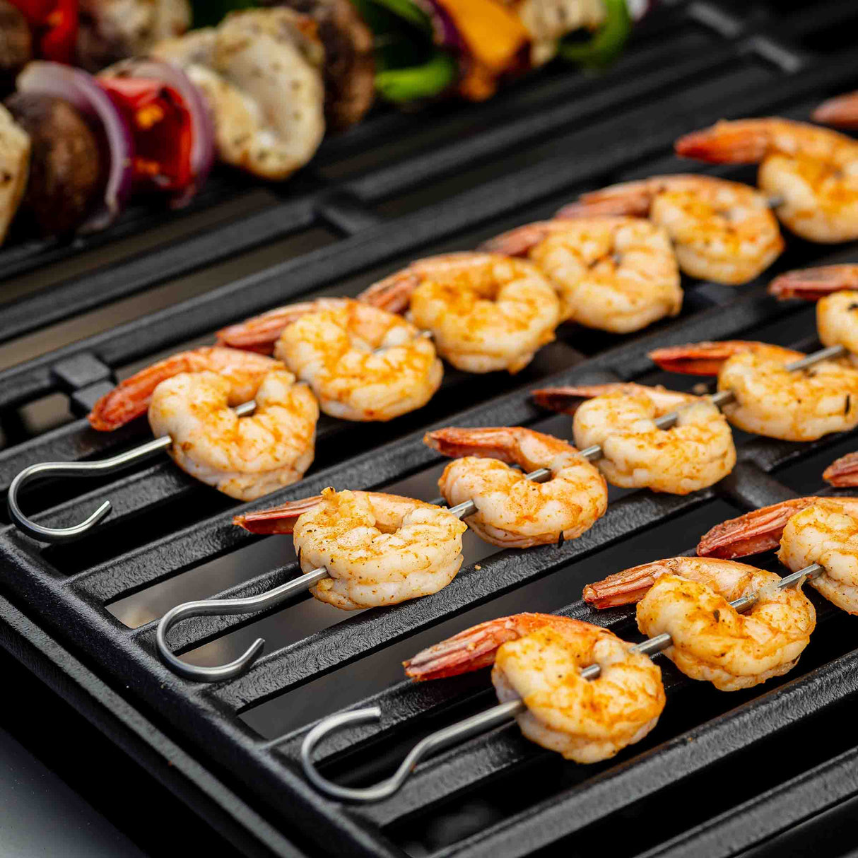 Rows of pink, seasoned shrimp are grilling on stainless steel skewers over a black grate. In the background, blurred vegetable and meat skewers hint at a variety of grilled dishes—all prepared using durable, dishwasher safe skewers.