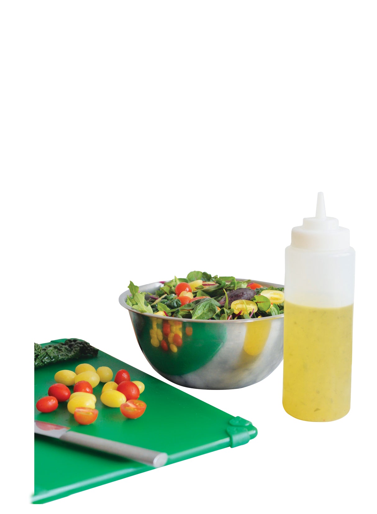 A green cutting board with a knife and sliced red and yellow cherry tomatoes sits next to a metal bowl of salad greens. A WideMouth Squeeze Bottle filled with yellow salad dressing stands upright beside the bowl on the white background.