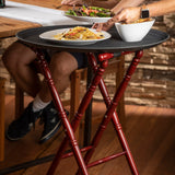 A person serves salad from a white bowl onto pasta on a round tray atop a slip-proof tray stand with a mahogany finish. Another person sits at a wooden table in the background, against a rustic brick wall and wooden floor.