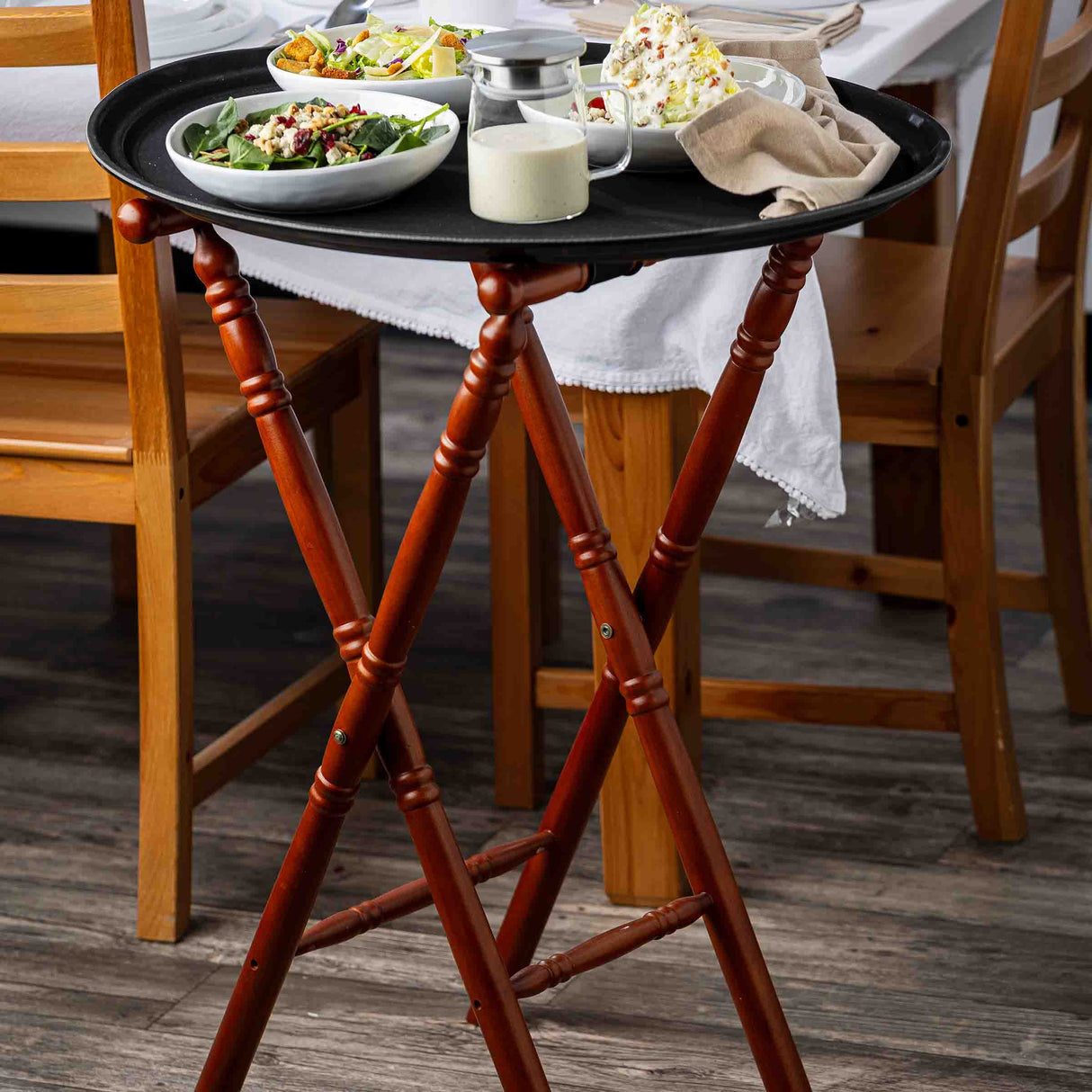 A wooden serving tray with a mahogany finish on a tray stand holds a glass jar of salad dressing, two small bowls of salad, and dessert with whipped cream. The tray sits beside a dining table set with white linens and chairs on a wood floor.