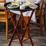 A wooden serving tray with a mahogany finish on a tray stand holds a glass jar of salad dressing, two small bowls of salad, and dessert with whipped cream. The tray sits beside a dining table set with white linens and chairs on a wood floor.