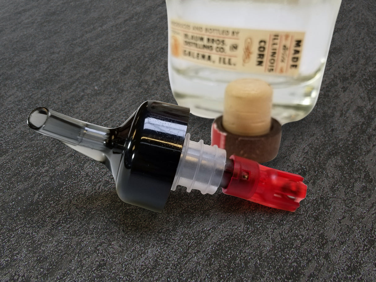 A close-up of a TableCraft Products Proper Pour 1 oz plastic pourer with a red base and black top lying on a gray textured surface. Behind it, a clear glass bottle with a wooden cork and label is slightly out of focus.