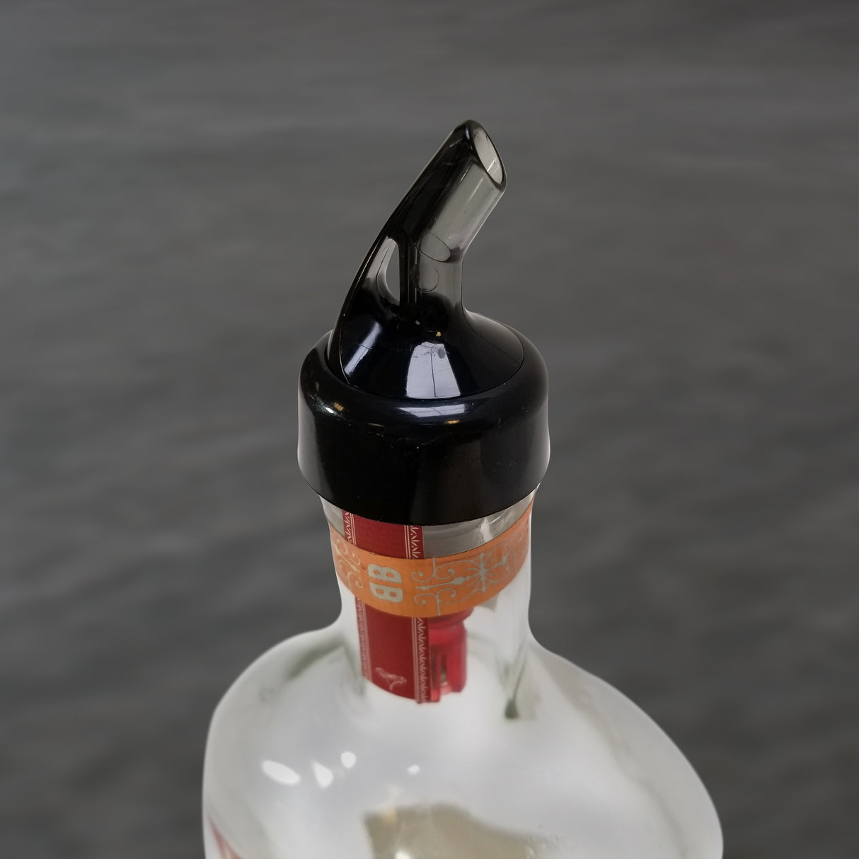 A close-up of a glass bottle with a TableCraft Products 1 oz plastic pourer on top. The bottle's brown and orange tax seal is partially visible, while the out-of-focus gray background highlights the Proper Pour spout and neck.