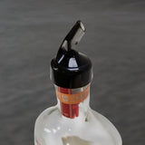 A close-up of a glass bottle with a TableCraft Products 1 oz plastic pourer on top. The bottle's brown and orange tax seal is partially visible, while the out-of-focus gray background highlights the Proper Pour spout and neck.