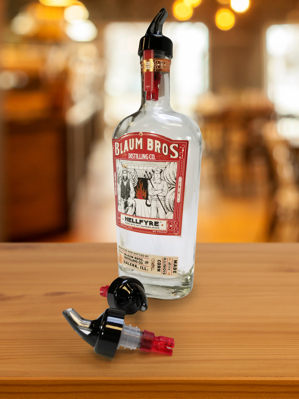 A clear bottle of Blaum Bros. Distilling Co. Hellfyre spirit with a red and white label sits on a wooden surface. A black 1 oz plastic pourer is attached, while another TableCraft Products pour spout lies nearby, suggesting a bar setting.