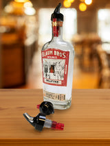 A clear bottle of Blaum Bros. Distilling Co. Hellfyre spirit with a red and white label sits on a wooden surface. A black 1 oz plastic pourer is attached, while another TableCraft Products pour spout lies nearby, suggesting a bar setting.