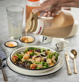 A hand pours creamy salad dressing from a large TableCraft bottle with an Option Dispenser Top onto a fresh salad of lettuce, cherry tomatoes, croutons, and chicken. Nearby are utensils, cups, and dishwasher safe items on a marble table.