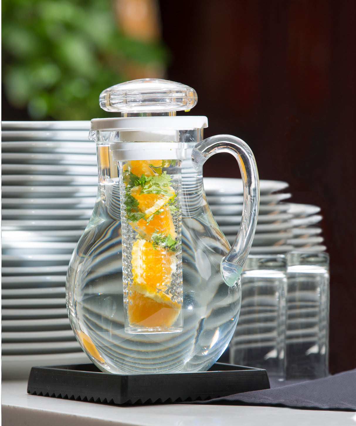A clear infused pitcher with a built-in infuser, filled with orange slices and mint, sits on a black tray. Stacked white plates and two empty glasses are behind it. The background is softly blurred, showcasing this fresh dishwasher safe pitcher.
