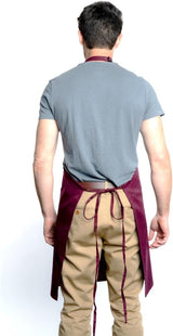 A man stands facing away, wearing a gray T-shirt, tan pants, and a burgundy chef approved bib apron with an adjustable neck tied at the back. His arms are relaxed by his sides against a plain white background, highlighting the apron’s back details and ties.