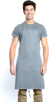 A smiling man with short dark hair wears a plain gray T-shirt, tan pants, and a chef approved bib apron with an adjustable neck and two front pockets. He stands facing forward against a white background, hands relaxed at his sides.