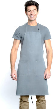 A smiling man with short dark hair wears a plain gray T-shirt, tan pants, and a chef approved bib apron with an adjustable neck and two front pockets. He stands facing forward against a white background, hands relaxed at his sides.