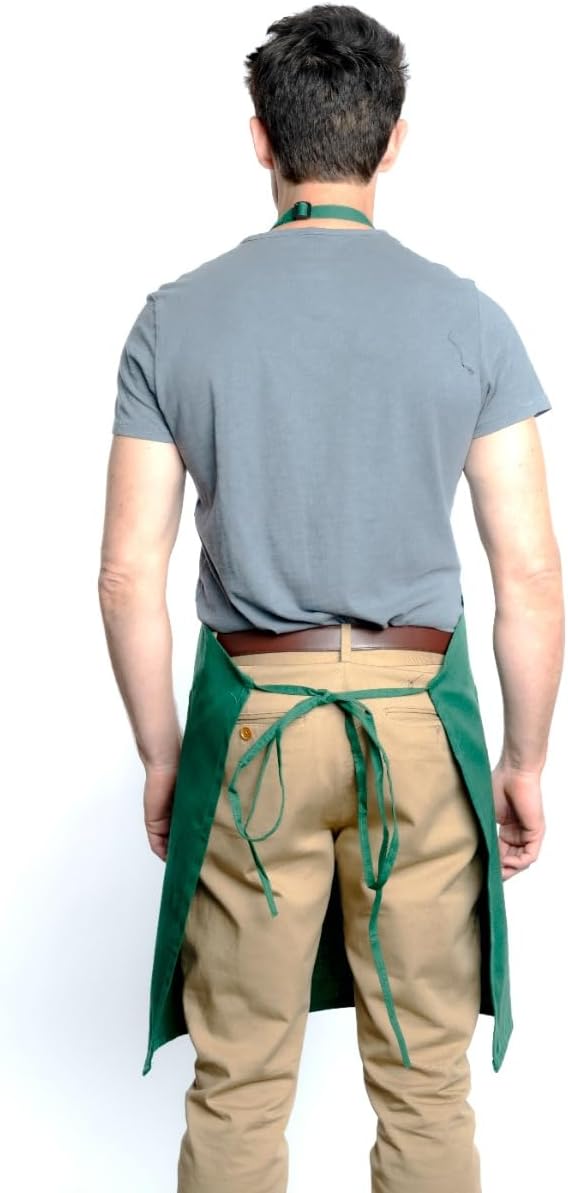 A man stands facing away, wearing a gray t-shirt, tan pants, and a green bib apron with an adjustable neck strap tied at the waist. The background is plain white, making this chef-approved look the main focus.