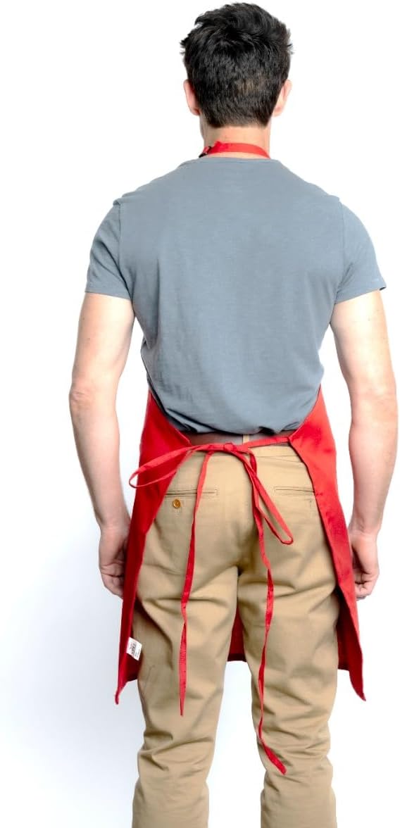 A man stands facing away, wearing a gray t-shirt, tan pants, and a chef-approved red bib apron with an adjustable neck tied at the back. The apron’s straps are visible as his arms hang relaxed at his sides. The background is plain white.