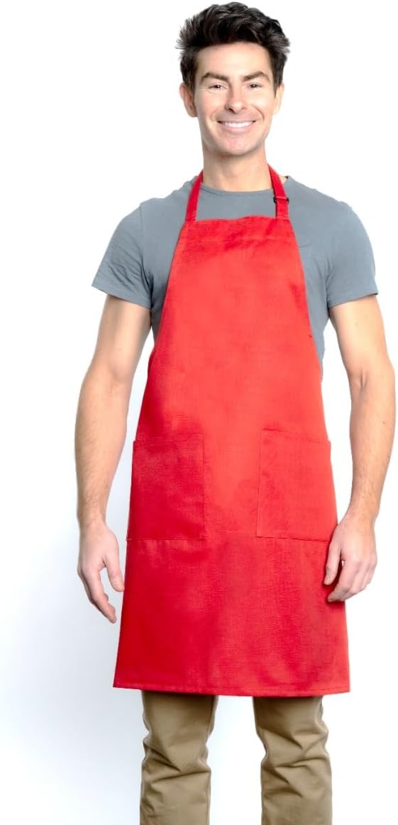 A smiling man with dark hair wears a gray t-shirt, tan pants, and a bright red chef approved bib apron with an adjustable neck and two front pockets. He stands facing forward against a plain white background.