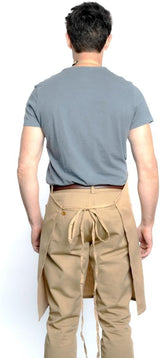 A person with short dark hair, wearing a light blue t-shirt and beige pants, is shown from behind. They have a chef approved beige bib apron with adjustable neck and long waist ties. The background is plain white.