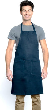 A smiling man wearing a grey t-shirt, tan pants, and a dark blue chef approved bib apron with an adjustable neck stands against a white background, facing forward with his arms relaxed at his sides. The apron has two pockets on the front.