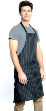 A man with short brown hair wears a gray t-shirt, light brown pants, and a chef approved black bib apron with thin white vertical stripes and an adjustable neck. He stands facing slightly right, smiling, against a plain white background.