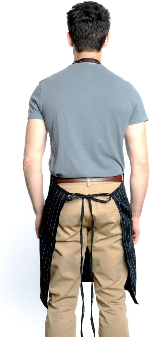 A man stands facing away from the camera wearing a gray t-shirt, tan pants, and a chef approved black pinstriped bib apron with an adjustable neck. His arms are relaxed at his sides, set against a plain white background.