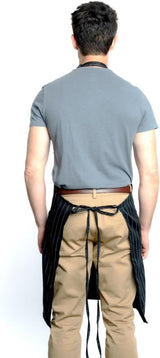 A man stands facing away from the camera wearing a gray t-shirt, tan pants, and a chef approved black pinstriped bib apron with an adjustable neck. His arms are relaxed at his sides, set against a plain white background.