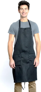 A smiling man stands against a white background, wearing a gray t-shirt, tan pants, and a chef approved bib apron with white vertical pinstripes. The apron features two front pockets and an adjustable neck strap. His arms are relaxed by his sides.