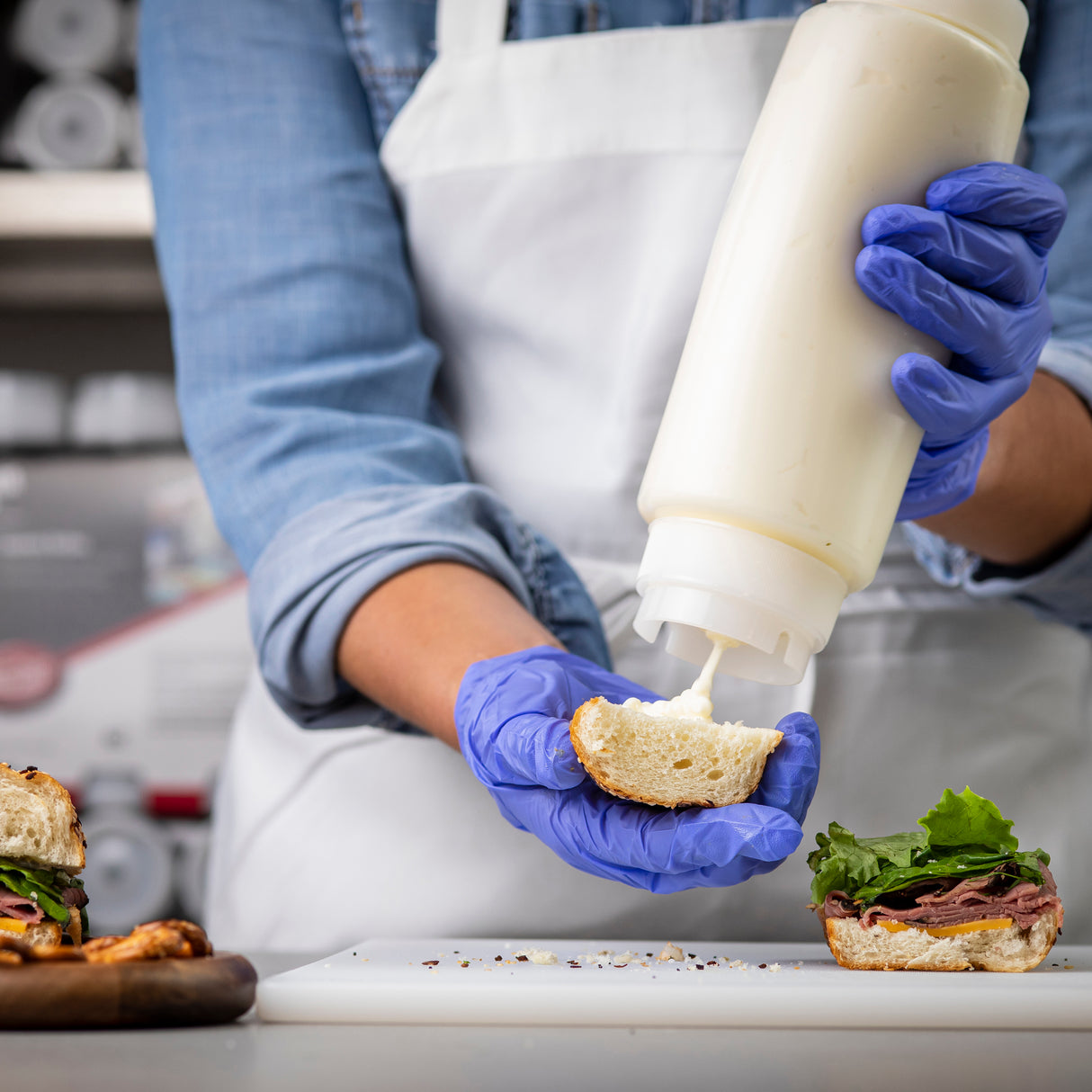 A person in a white apron, blue denim shirt, and blue gloves uses a squeeze dispenser to add mayonnaise onto a sandwich bun over a cutting board. An open-faced sandwich with lettuce and meat is nearby in the kitchen.