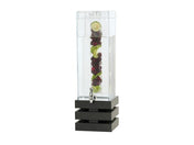 A tall, clear BPA free beverage dispenser with a square black base is filled with water. Inside, a vertical infusion chamber holds lime slices, cherries, and green herbs. A silver spigot is positioned near the bottom for dispensing drinks.