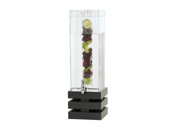 A tall, clear BPA free beverage dispenser with a square black base is filled with water. Inside, a vertical infusion chamber holds lime slices, cherries, and green herbs. A silver spigot is positioned near the bottom for dispensing drinks.