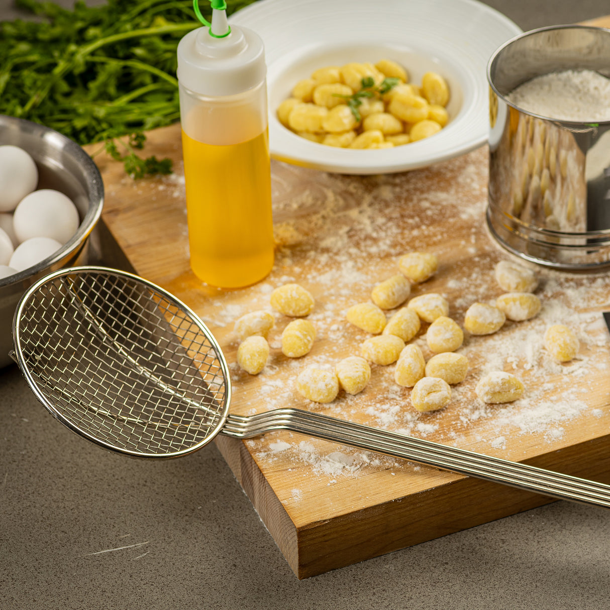 A nickel-plated skimmer with a square mesh rests on a floured wooden board with uncooked gnocchi, surrounded by a bowl of eggs, cooked gnocchi, oil, fresh herbs, and a sifter dusted with flour on the kitchen counter.