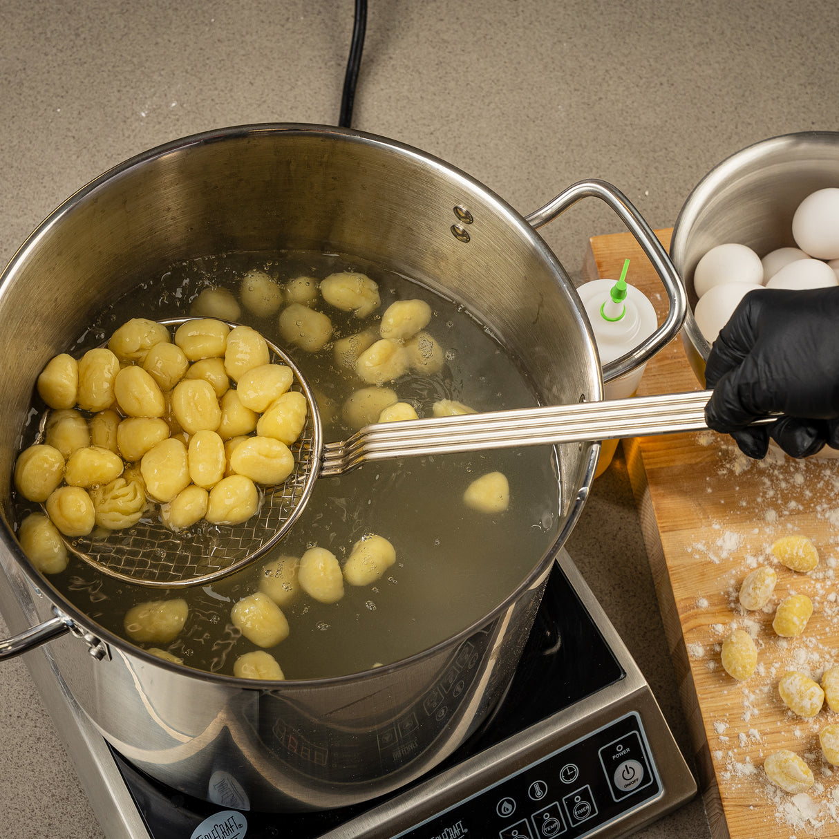 A hand wearing a black glove uses a nickel plated skimmer to lift cooked gnocchi from a pot of boiling water on an induction cooktop. Uncooked gnocchi, eggs, and flour rest on a wooden board nearby. A squeeze bottle and countertop are also visible.