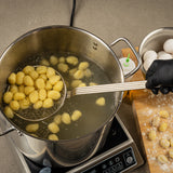 A hand wearing a black glove uses a nickel plated skimmer to lift cooked gnocchi from a pot of boiling water on an induction cooktop. Uncooked gnocchi, eggs, and flour rest on a wooden board nearby. A squeeze bottle and countertop are also visible.