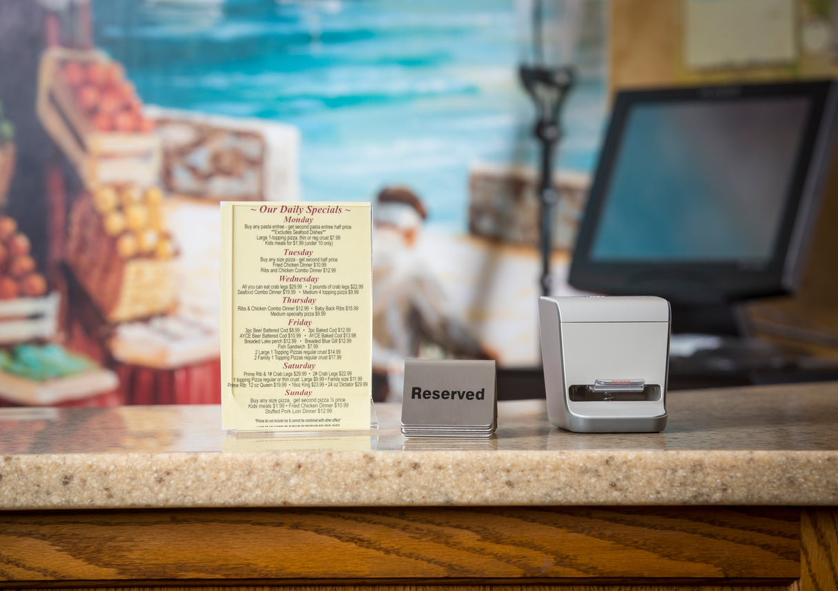 A restaurant counter displays a printed menu, a metal “Reserved” sign, and a TableCraft Products toothpick dispenser next to a white card holder. In the background, there’s a blurred mural of a market scene and a computer monitor.